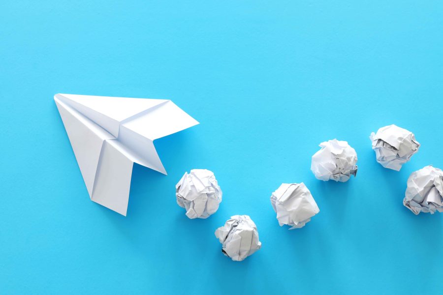 A white paper airplane flying ahead, while a series of crumpled paper balls trail behind on a blue background.